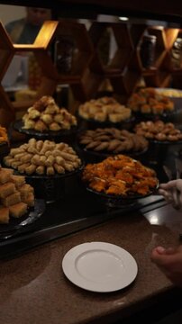Vertical screen hand in gloves serves golden basbousa from a rich display of Middle Eastern sweets stacked in generous piles under warm lights creating a cozy indulgent dessert moment