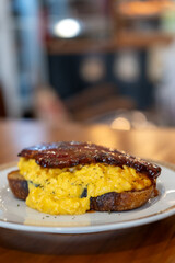 Toast topped with scrambled eggs and thick cut bacon, served on a plate 