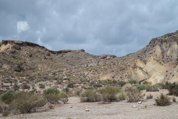 A rugged desert landscape in Tabernas, Almería, featuring arid hills, sparse vegetation, and dry terrain under a cloudy sky. The scenery evokes a cinematic, wild western atmosphere