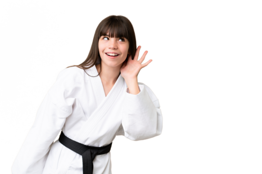 Little Caucasian girl doing karate over isolated background listening to something by putting hand on the ear
