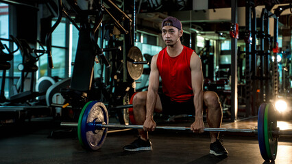 Obraz premium Young Asian man in red sleeveless shirt weightlifting with barbell in modern gym, demonstrating strength, fitness, and focused workout routine