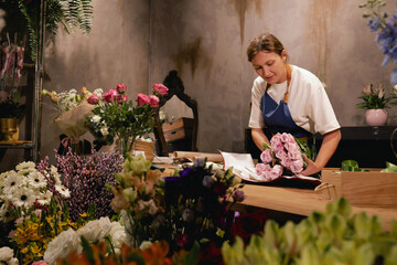 Middle-aged female florist in apron arranging fresh flowers for bouquet in the flower shop. Small...