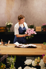 Middle-aged female florist in apron arranging fresh flowers for bouquet in the flower shop. Small...