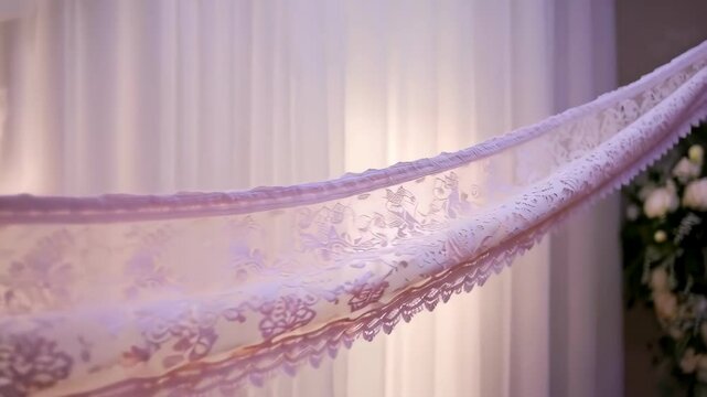 Close-up of delicate lace fabric with floral patterns and scalloped edges suspended against a soft white draped curtain backdrop.