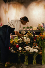 Middle-aged female florist in apron arranging fresh flowers for bouquet in the flower shop. Small...