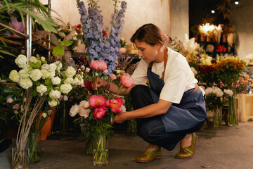 Middle-aged female florist in apron arranging fresh flowers for bouquet in the flower shop. Small...