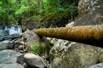 Rusty metal pipe with lichen and corrosion over mossy rocks in a natural outdoor setting, showing weathered textures and the contrast between nature and decay.