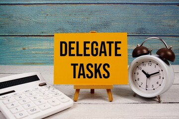 Delegate Tasks text written on paper card with calculator and alarm clock on wooden background