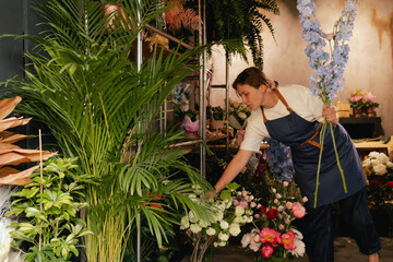 Middle-aged female florist in apron arranging fresh flowers for bouquet in the flower shop. Small...