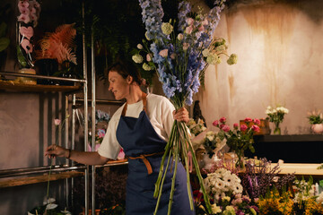 Middle-aged female florist in apron arranging fresh flowers for bouquet in the flower shop. Small...