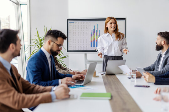 Female executive director standing at boardroom with her team of experts around conference table, discussing and analyzing data.