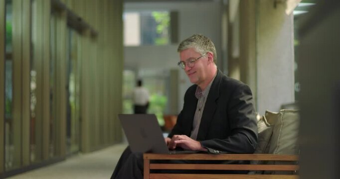Middle aged man smiling slightly while working on laptop in relaxed office setting, seated comfortably in modern corridor workspace