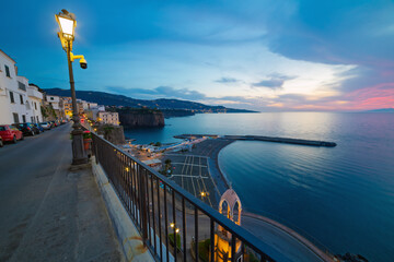 Sunset view Meta di Sorrento, Italy. Coastal town illuminated by street light. Calm sea water, beautiful sky with clouds. Mountains on horizon.
