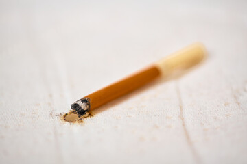 Close-up of cigarette resting against white fabric, leaving burn mark. Soft lighting highlights damage, contrasting with brown and white tones. Shallow depth of field focuses on burn area
