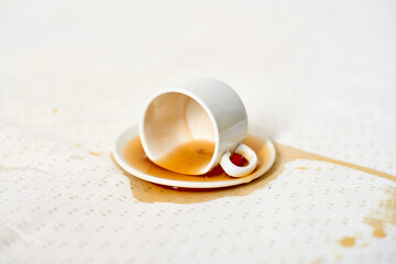 White cup, tipped on side, rests on saucer, with spilled coffee staining fabric. Soft lighting highlights spill pattern on cloth surface, creating subtle contrast