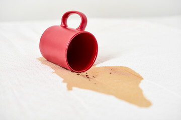 Red mug tips over, spilling coffee on white surface. Brown liquid stain spreads, creating contrast...