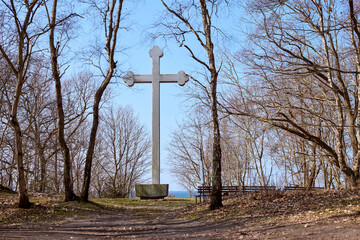 Tall cross stands prominently in leafless forest clearing. Clear blue sky serves as backdrop, enhancing tranquility. Bare trees frame path leading to cross
