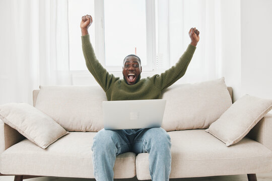 Excited young Black man celebrating success while working on a laptop in a cozy living room Happiness, motivation, and productivity are emphasized