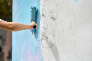 Hand applies soft blue paint over pencil sketch of young female face on white wall. Natural outdoor setting, gentle lighting, artistic enhancement in progress © TRAVELARIUM
