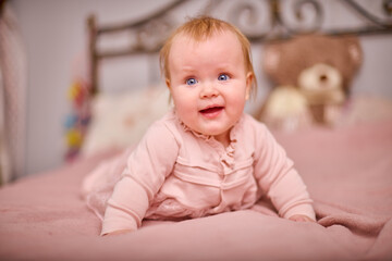 Naklejka premium Young female with blonde hair, light skin, smiles joyfully while lying on pink bed. Soft lighting and ornate headboard create cozy, warm atmosphere