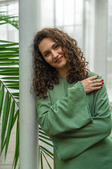 Charming young woman in green dress leaned gently against white pillar. In background are green...