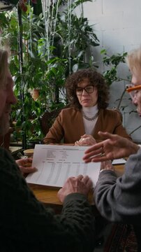 Medium vertical shot of professional young Caucasian female financial adviser consulting client couple on terms of bank loans, demonstrating document with amortization schedule and interest rates