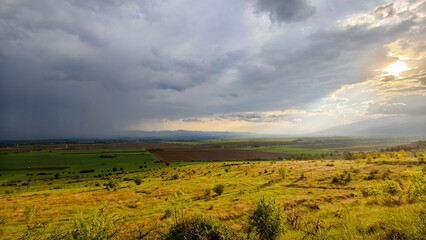 A dramatic rural scene where a downpour drenches one side of the field, while sunlight shines...