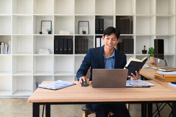 Smiling businessman in a modern office uses a laptop while referencing notes in a notebook. A productive work session surrounded by charts, documents, and a clean organized workspace