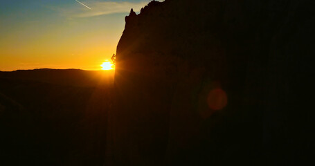 A person is rock climbing on a steep cliff as the sun sets in the background. The silhouette of the...
