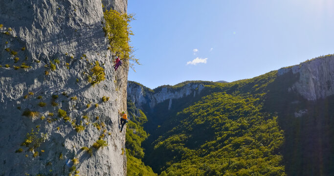 A breathtaking aerial view showcases two climbers scaling a steep rock face, surrounded by lush green mountains and a clear blue sky. The climbers' determination and skill are evident as they conquer.