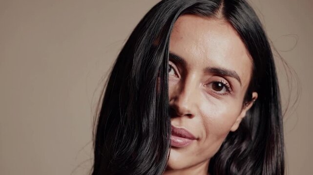 Close-up video shot of a woman with long dark hair against a neutral background, capturing a serene expression from a slightly tilted angle.