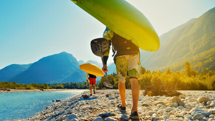 Two men, one of whom is of Asian descent, walk along a rocky riverbank carrying surfboards. The scene is a close-up shot.