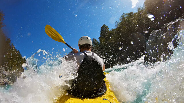 A thrilling kayaker, donned in a white helmet and black life jacket, navigates a yellow kayak through a turbulent river. The camera captures a close-up shot from behind.