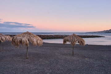 Tropical beach with straw umbrellas at sunset, volcanic sand shore