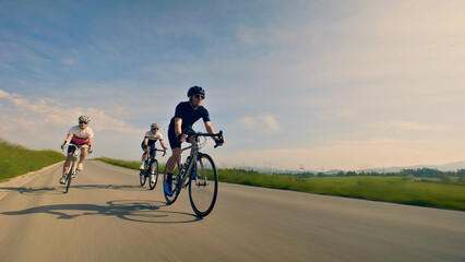 Obraz premium Three helmeted cyclists pedal down a scenic country road, with a man in a black shirt and sunglasses leading the pack. The camera captures a dynamic medium shot.