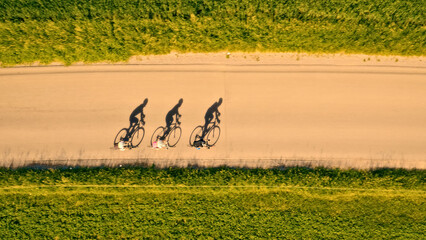 An aerial view captures three cyclists riding in a line on a dirt road, surrounded by lush greenery. The silhouettes of the riders and their bikes are visible. © wkproduction