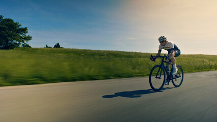 A man, dressed in cycling gear and wearing a helmet, rides a bicycle on a paved road. The camera captures a dynamic, fast-paced shot, emphasizing the cyclist's speed and the surrounding greenery. - obrazy, fototapety, plakaty