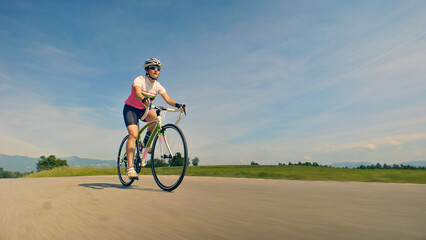 A woman, clad in a pink and white shirt and helmet, rides her bicycle on an open road. The shot is a dynamic medium shot, emphasizing her speed and the vast, scenic landscape around her.