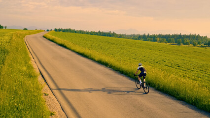 A person rides a bicycle down a winding road, surrounded by lush green fields. The shot is an aerial view, showcasing the cyclist's journey through the picturesque countryside. - obrazy, fototapety, plakaty