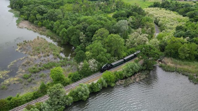 Steam train engine locomotive arriving and slowin down to lakeside station near Lednice, Czech Republic. Aerial drone camera reverse to follow train speed. Green trees and lake water reflection.