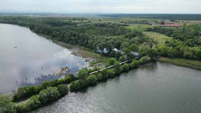 Steam engine train departing lakeside station near Lednice, South Moravia, Czech Republic. Aerial drone spring green trees view. Wide angle camera follows the train movement.
