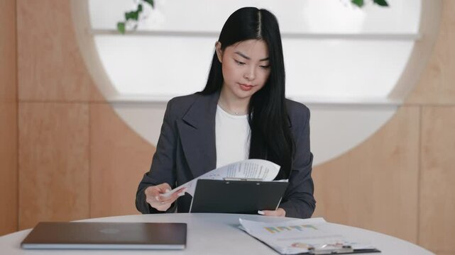 Determined Asian office worker flipping through documents clipped to folder while sitting at desk. Reviewing charts and business data attentively while analyzing reports or preparing meeting notes.