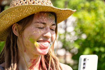 Woman in Straw Hat with Colorful Powder on Face and Dreadlocks Holding Mobile Phone for Selfie, Celebrating Holi.