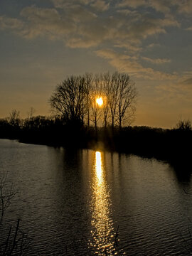 Silhouettes of trees and lake with reflections in the rippling watr of a cloudy evening sky with golden sun glow in the valley of ude Kale river, Vinderhoute, Flanders, Belgium
