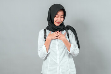 Obraz premium Modest Muslim student wearing a hijab and backpack, smiling gratefully with hands on chest. Back to school concept, expressing humility and enthusiasm in a studio shot.