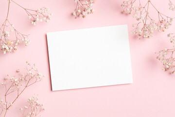 Horizontal white blank card mockup framed by baby breath flowers on soft pink background, ideal for wedding invitations or greeting cards, flat lay