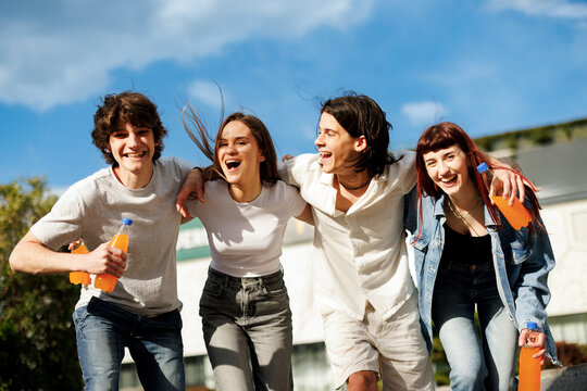 Four students are running and laughing together outside, holding orange drinks on a sunny day