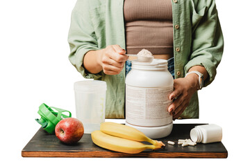 Woman preparing protein drink, holding measuring spoon with whey protein powder above plastic jar on wooden board with white capsules, shaker, bananas and apple. Isolated on a transparent background