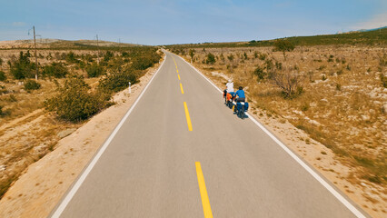 A group of cyclists, including a man in a blue shirt, ride their bikes down a long, empty road in a desert-like area. The aerial view showcases their journey through the arid landscape.