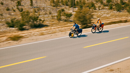 Obraz premium Aerial view: Two cyclists ride their loaded bikes down a highway, showcasing their cross-country travel and bike touring adventure. The person in the front sports a blue plaid shirt.